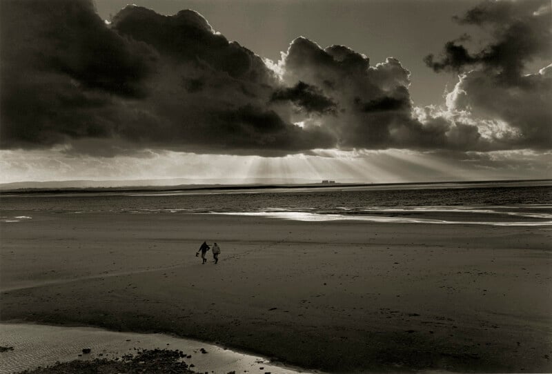 Two people walk hand in hand across a vast, empty beach under dramatic clouds, with sunlight streaming through gaps, illuminating the sand and distant water.
