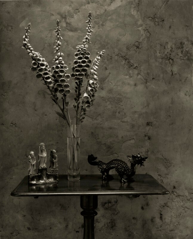 A black-and-white photo of a small wooden table with a glass vase of tall foxglove flowers, three small figurines of people, and a decorative dragon sculpture against a textured wall.