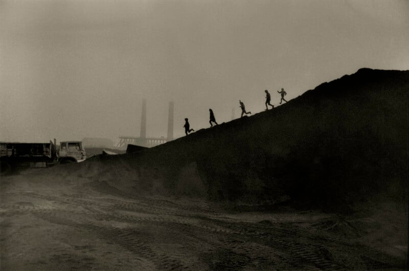 Silhouetted children run up a steep dirt mound near industrial chimneys, with a truck and vehicle tracks in the foreground, under a hazy sky.