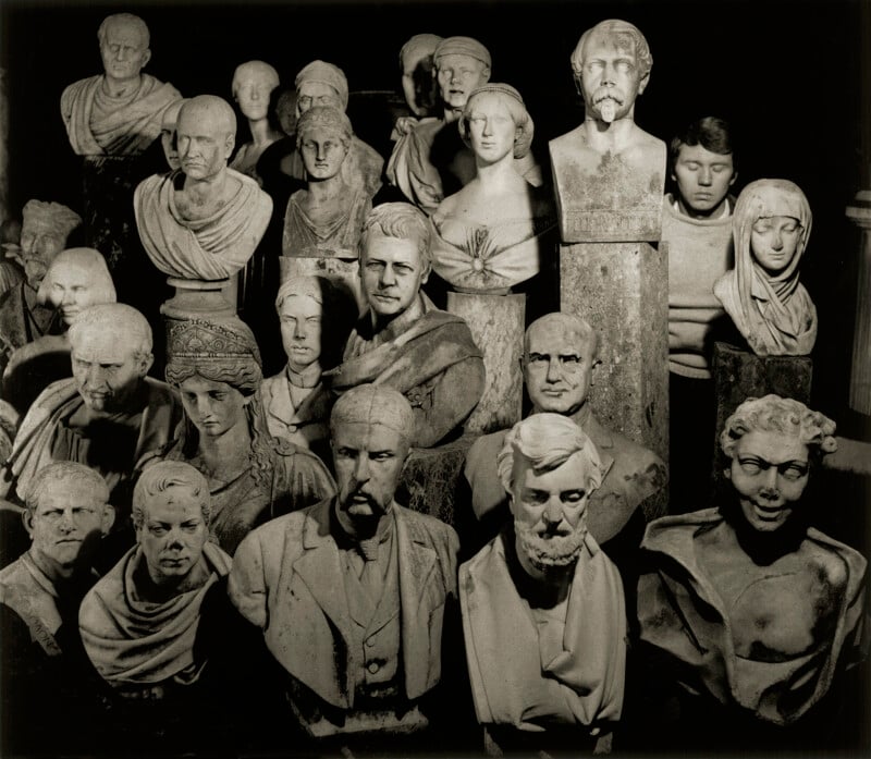 A group of various marble and plaster busts arranged closely together, depicting men and women with different facial expressions, hairstyles, and clothing styles, all lit dramatically against a dark background.