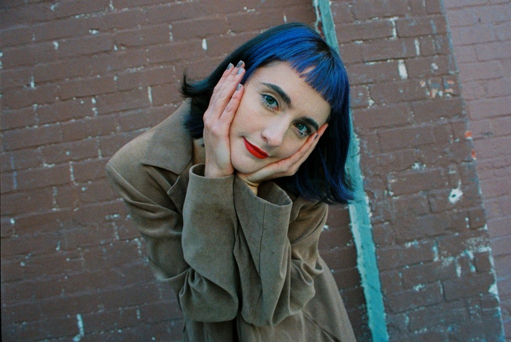 A smiling female singer with blue hair with her head in her hands.
