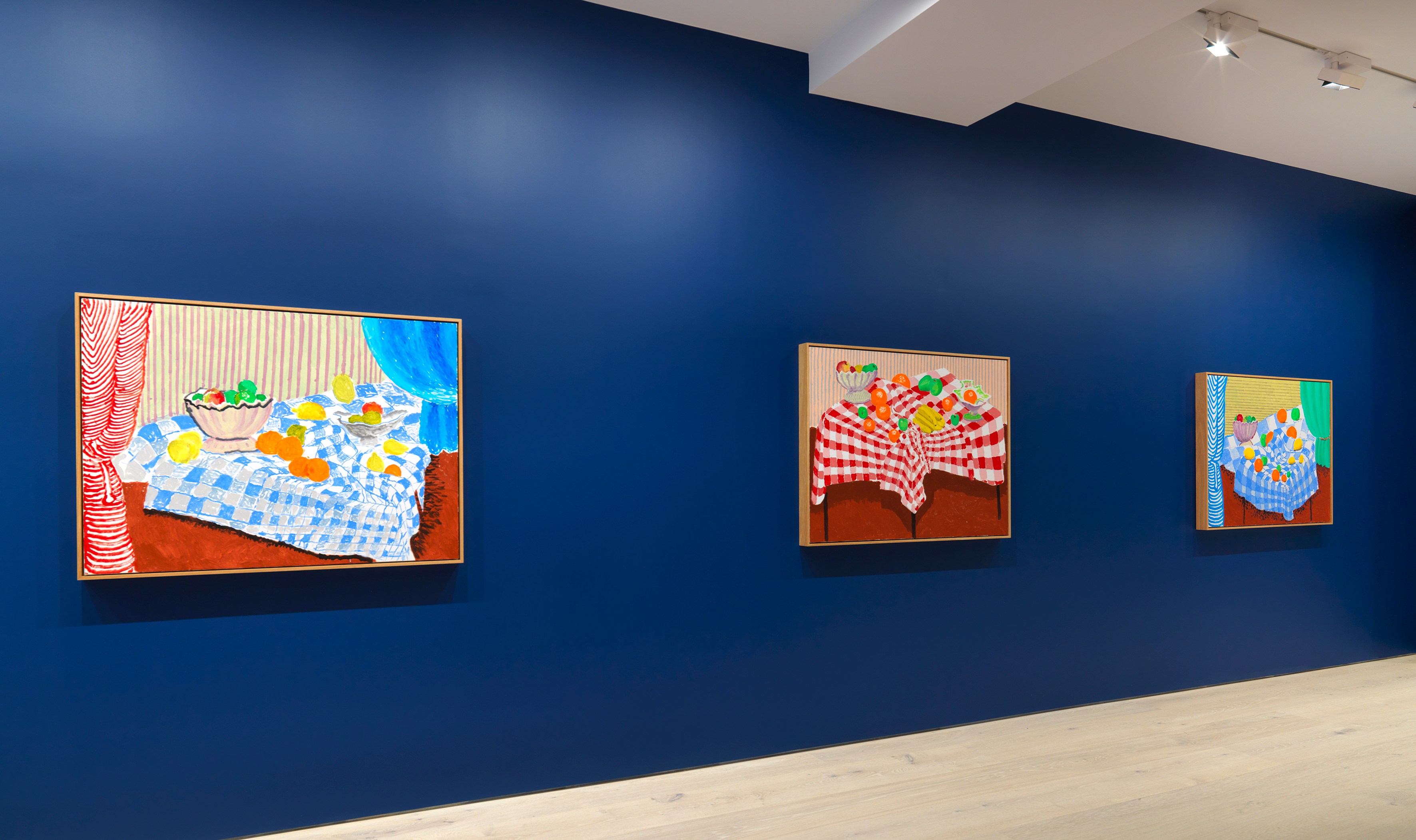 Installation view of three David Hockney still life paintings on a dark blue wall.