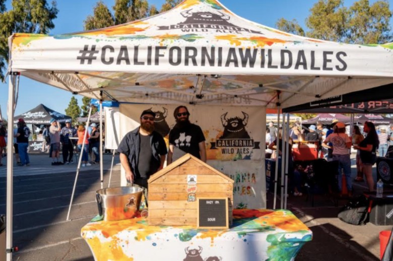 Two men stand under a canopy bearing the label #californiawildales as part of the Capital of Craft Beer Fest.