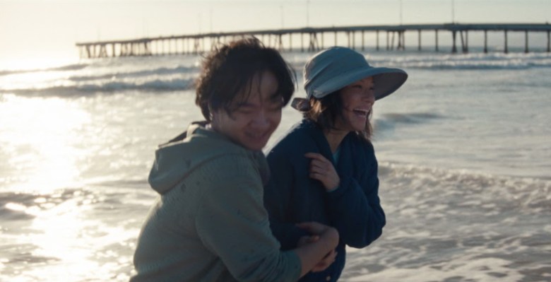 A man and woman embrace and laugh on a beach with the sun shining over the water and a pier visible behind them. It's a still from the film "Rosemead," featured in the San Diego Asian Film Festival.