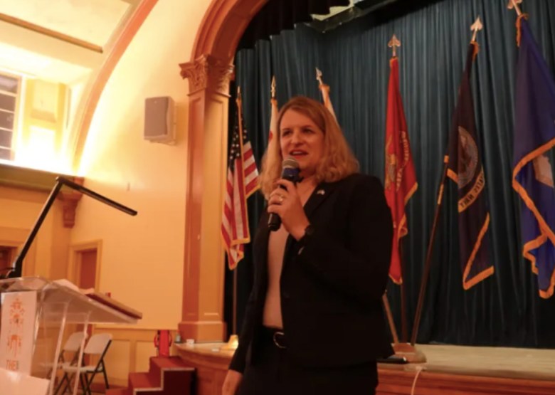 A woman stands in front of a stage, holding a microphone in a community hall. Its a veterans event.