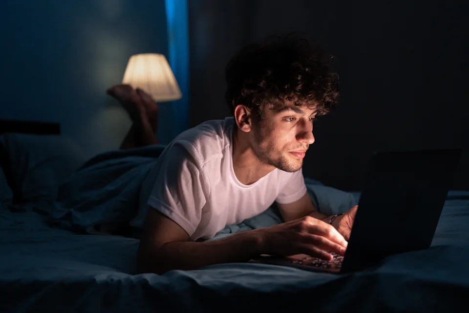 Person lying on a bed, focused on a laptop screen, with a lamp softly illuminating the room