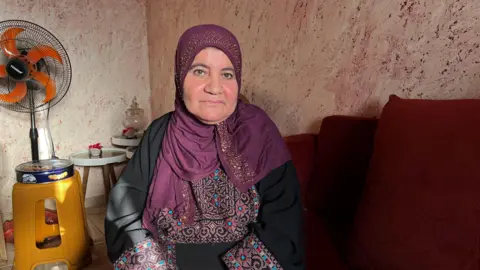 Afaf abu Alia. She is wearing a black dress with patterns around the sleeves and on the chest. She is wearing a deep red heascarf with gold detailing. She is sitting on a red sofa, the wall behind her is beige with a red pattern and there is a fan next to some tables nearby
