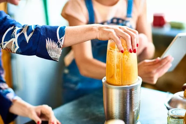 Hand of woman preparing smoothie in blender, daughter holding tablet in background