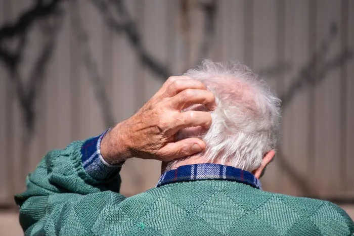 Elderly person scratching head, wearing a quilted jacket and a plaid shirt, viewed from behind
