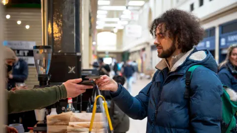 Getty Images A young man with curly hair and a blue puffer jacket paying for something at a coffee shop counter using his bank card 