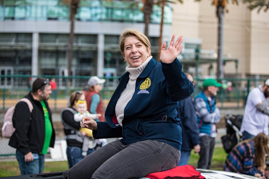 A person, with short blonde hair, smiles and waves to a crowd of attendees while sitting on top of a car during a parade. The person wears a beige turtle neck with a blue jacket.