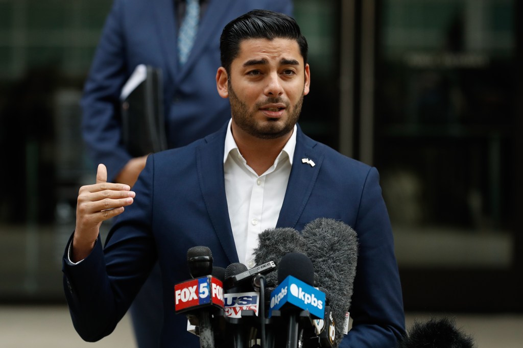 A person — with slick-back dark hair and wearing a blue suit — stands in front of stand with several microphones on it as they speak during a press conference in front of a courthouse.