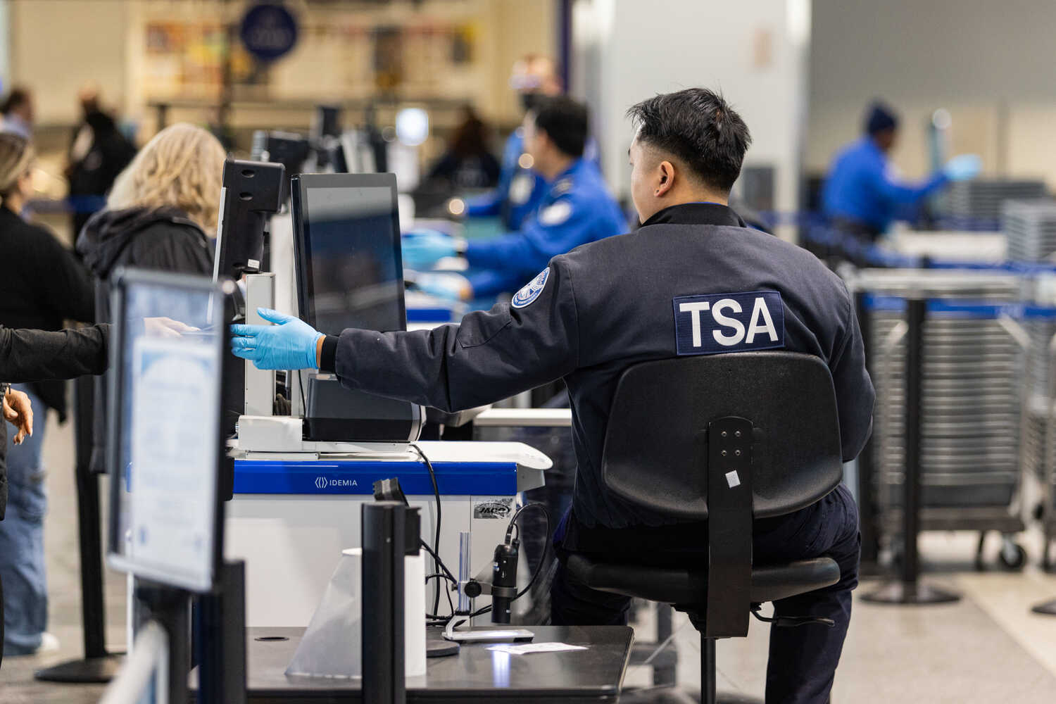 A person with a blue jacket that says TSA sits behind a computer.