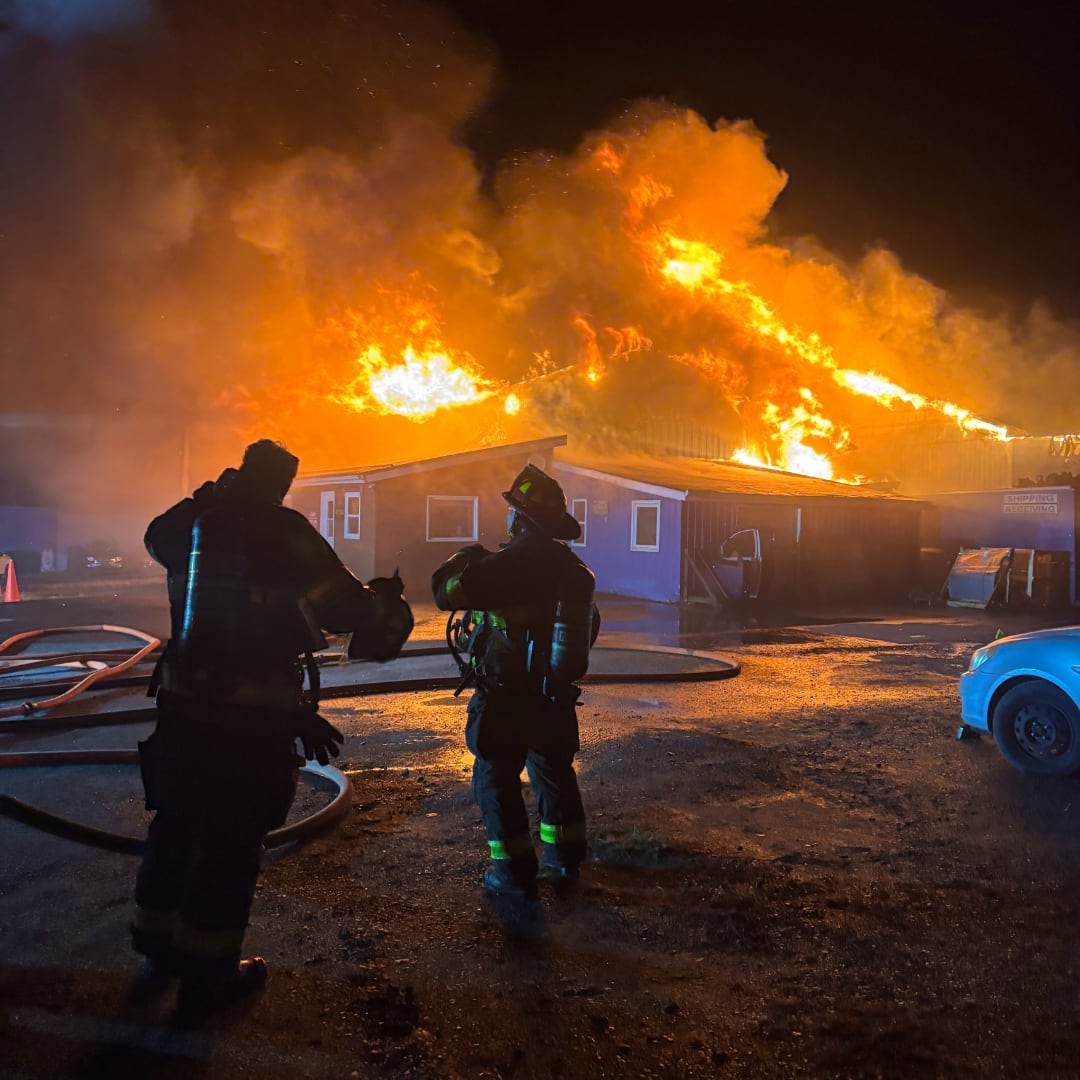 A building on fire, large flames coming out of the roof, two firefighter's standing in front. 