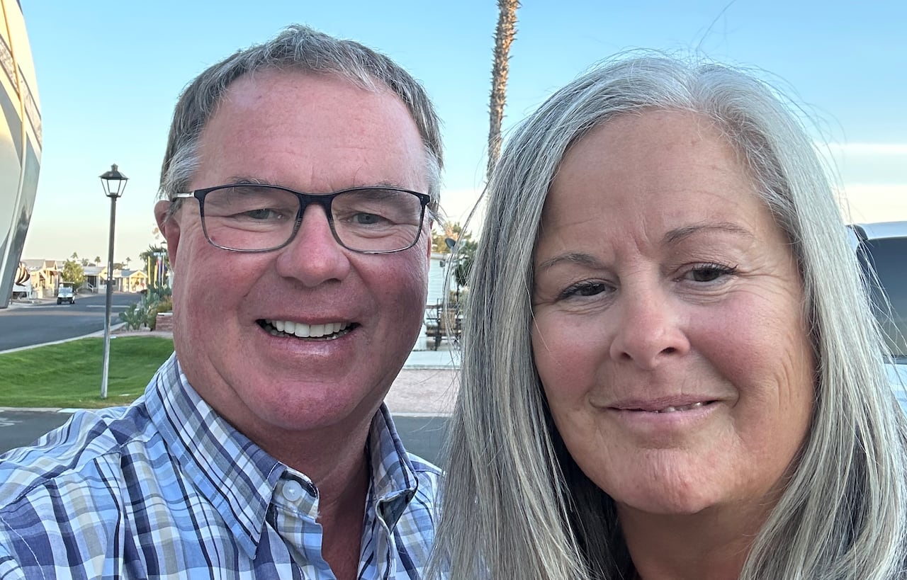 A man and a woman with greying hair pose for a photo outside in front of a palm tree.