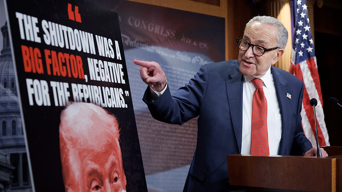 Senate Minority Leader Chuck Schumer stands at a podium and points at a poster of Donald Trump with a quote.