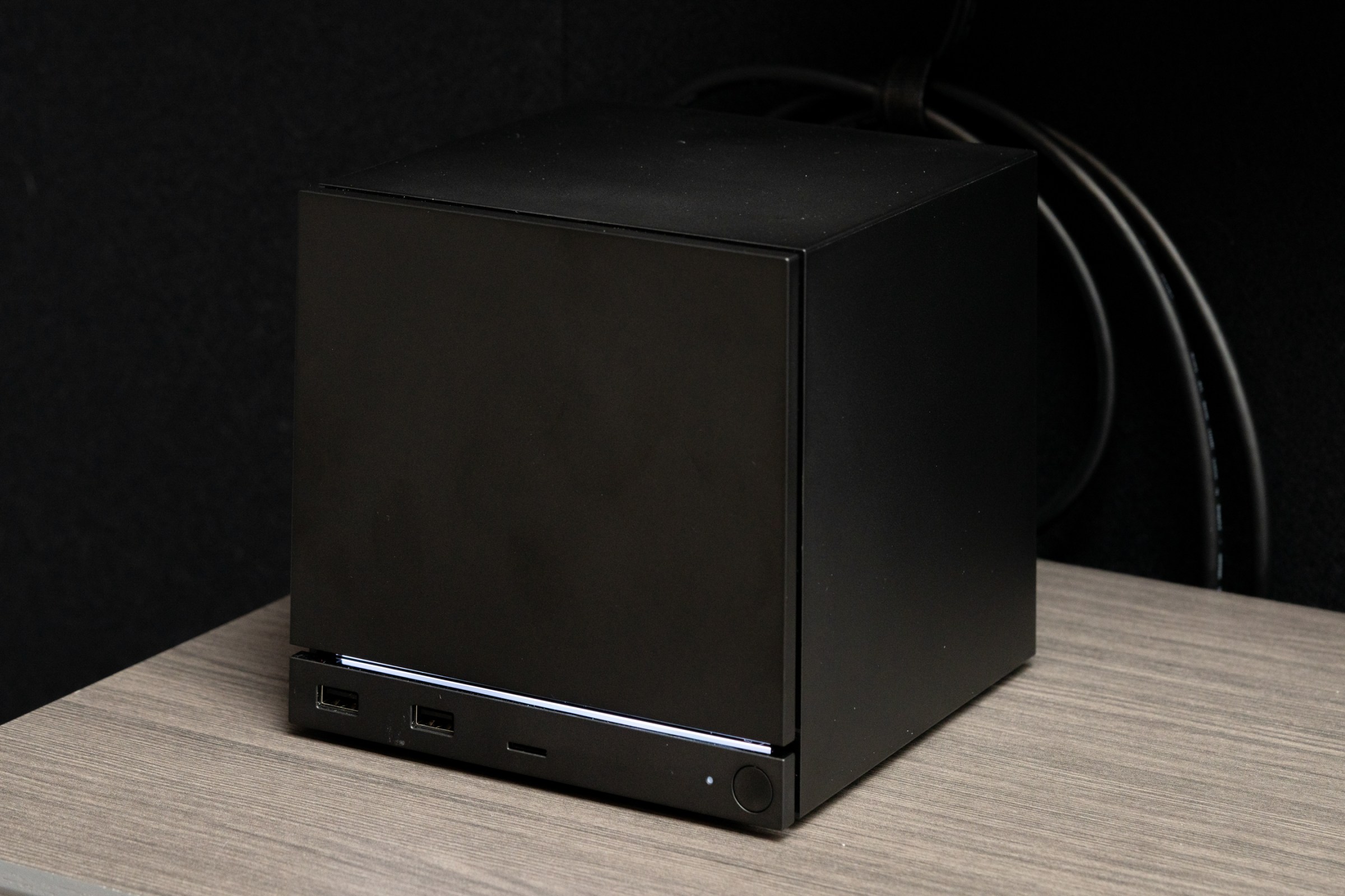 The Steam Machine on a gray wooden table, up close, in the shadows of a TV, showing cords plugged in and the light bar lit up.