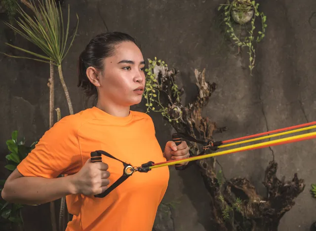 A twenty-something woman does standing back rows with a resistance band. Working our and training at a home gym.