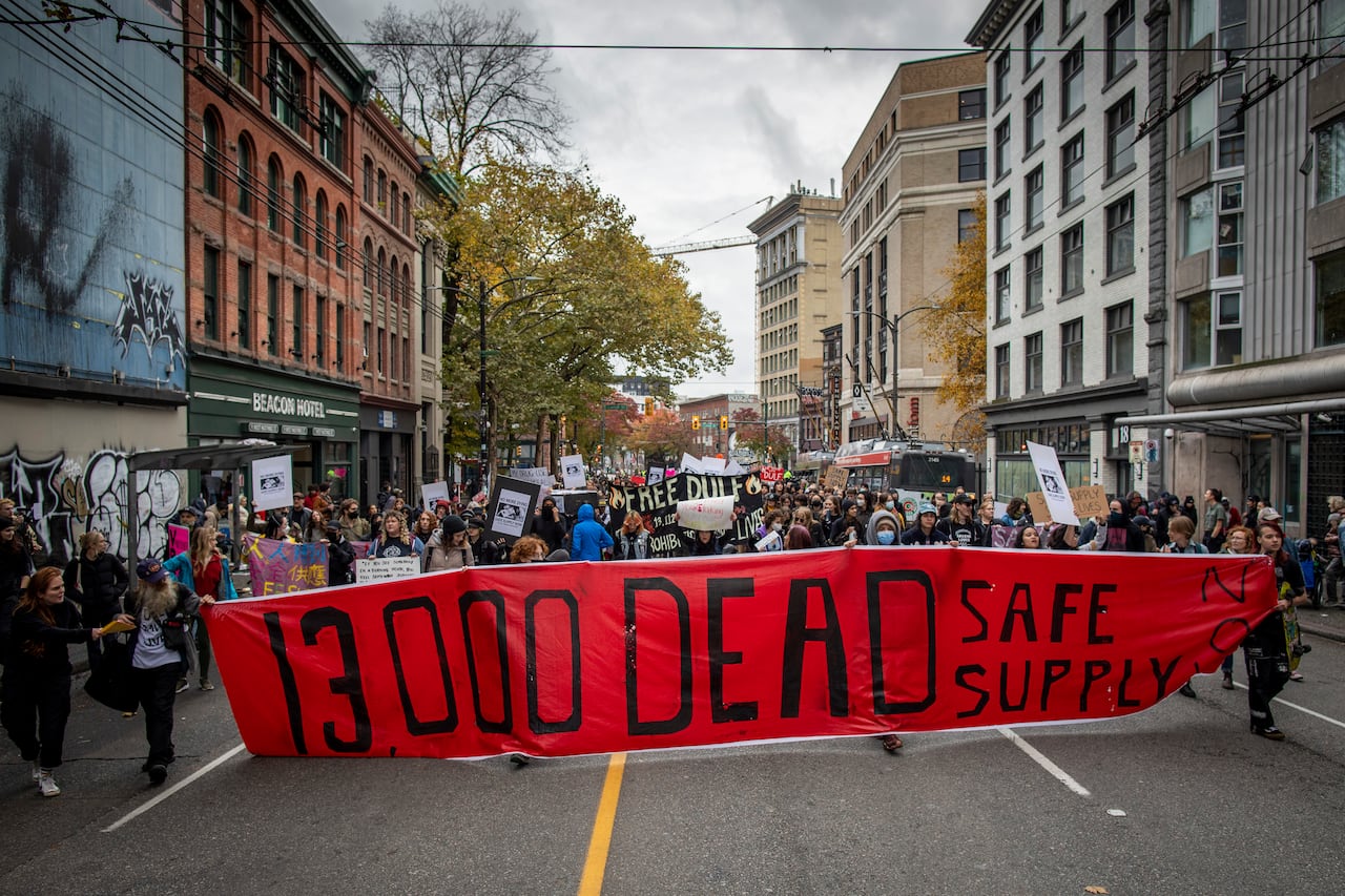 A crowd carries a large banner that says "13,000 dead, safe supply now."
