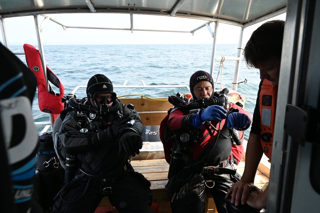 Two men in scuba gear sit in the back of a boat as they pull on gloves and check respirators on the sparkling blue waters of Lake Ontario. 
