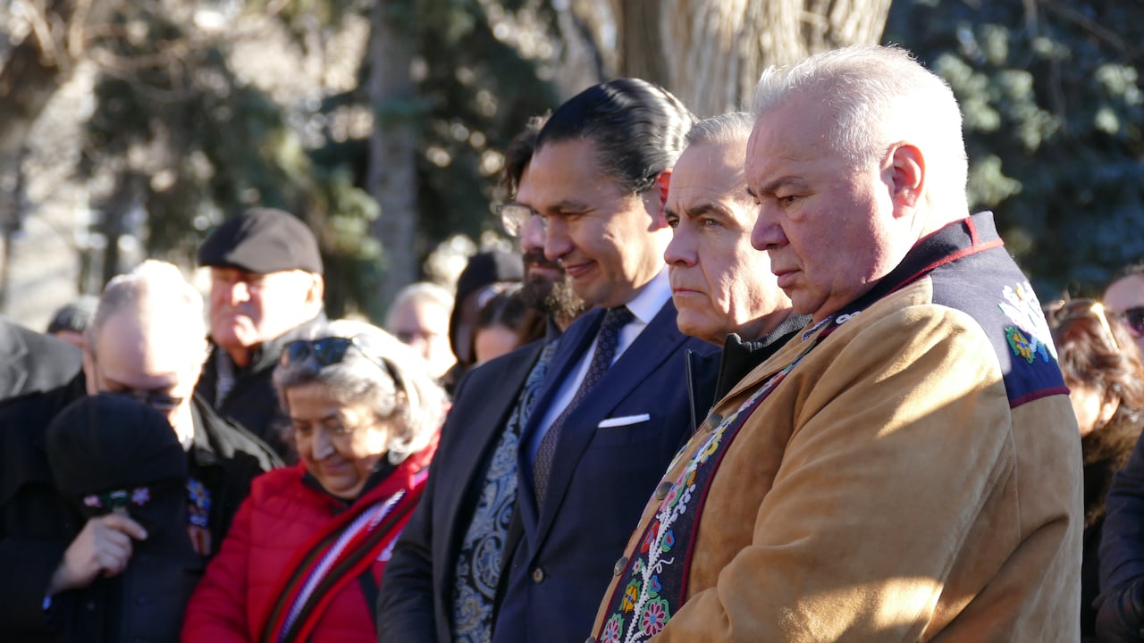 Man wearing light brown jacket with beaded floral design stands next to two men in suits among a crowd