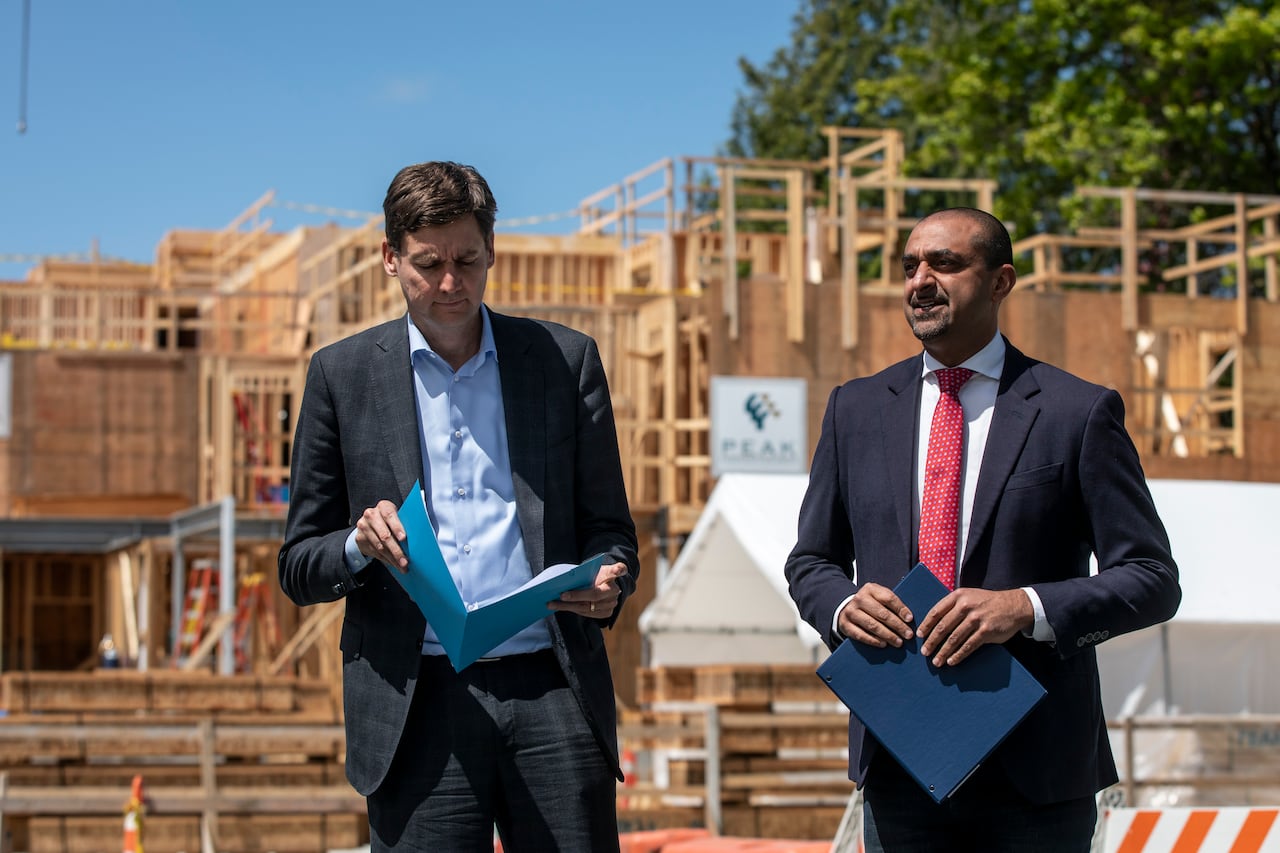 Two tall men are pictured holding files while standing in front of an under-construction home.