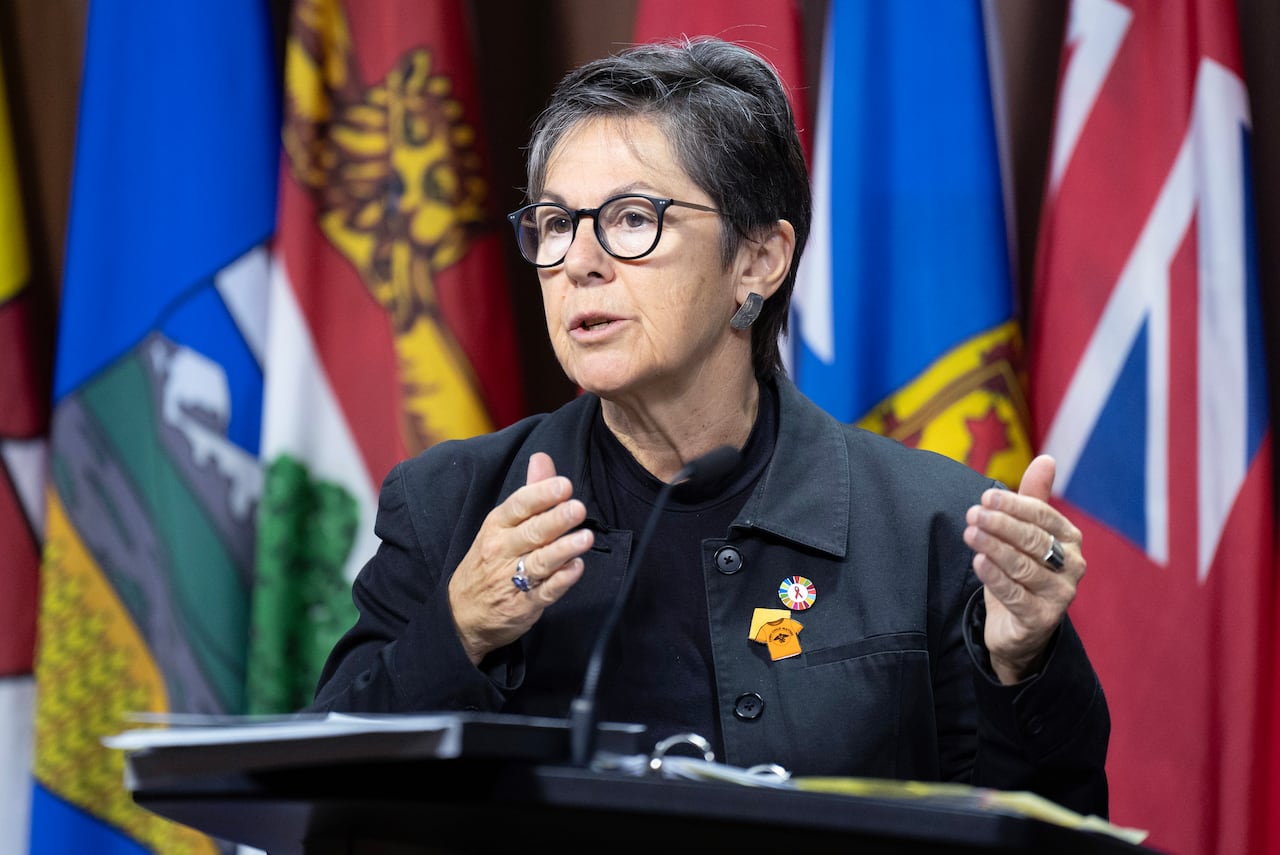 A woman speaks at a podium, with flags behind her.