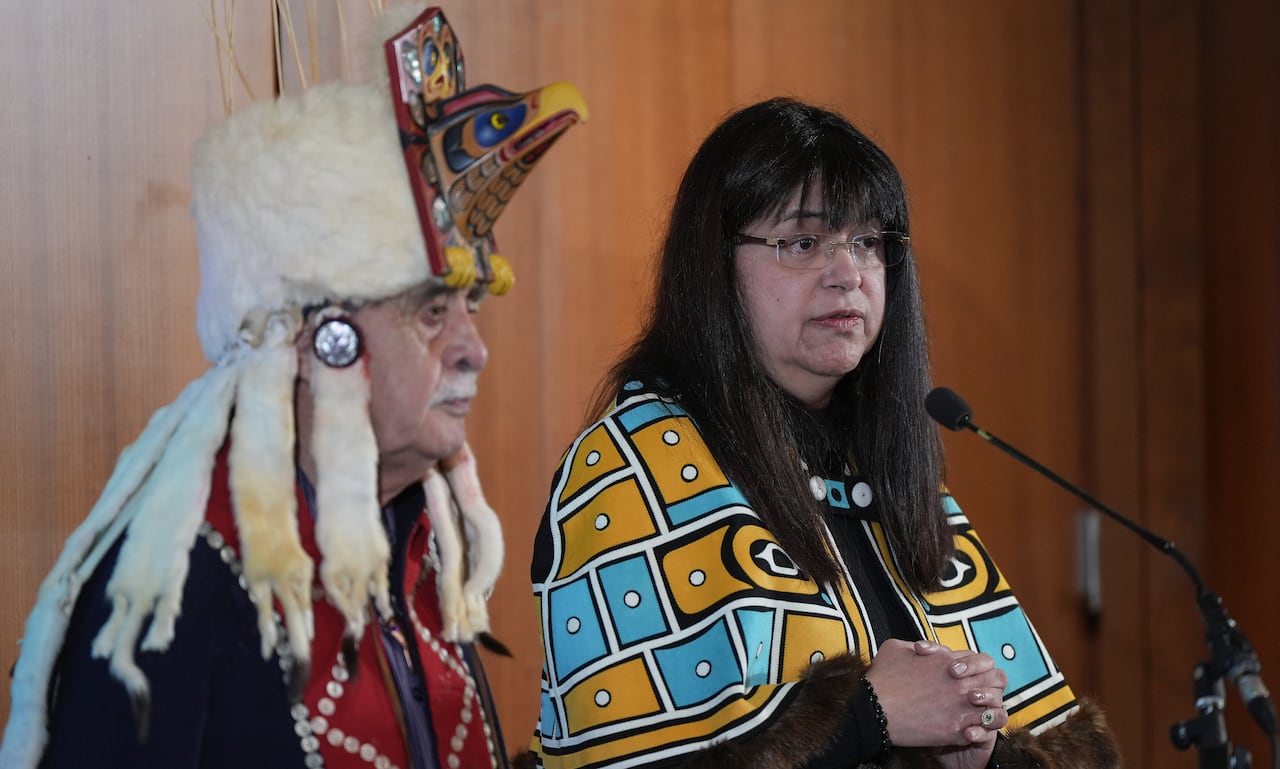 A man and a woman wearing Indigenous regalia speak to a mic.