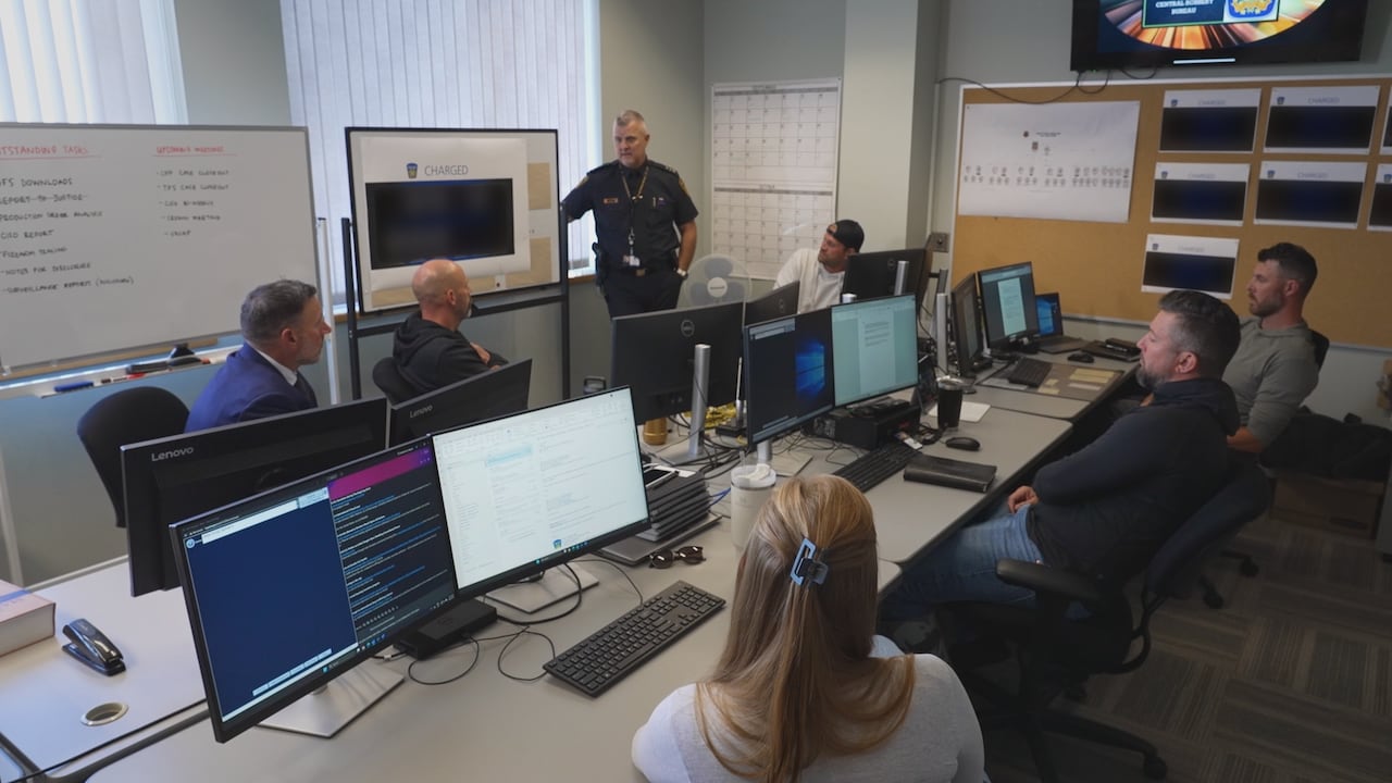 Police sit in a meeting nearby their office workstations, focusing on one uniformed officer at a whiteboard.