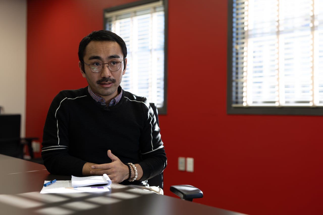 A man sits at a boardroom table with notes in front of him, a bright red wall behind him.