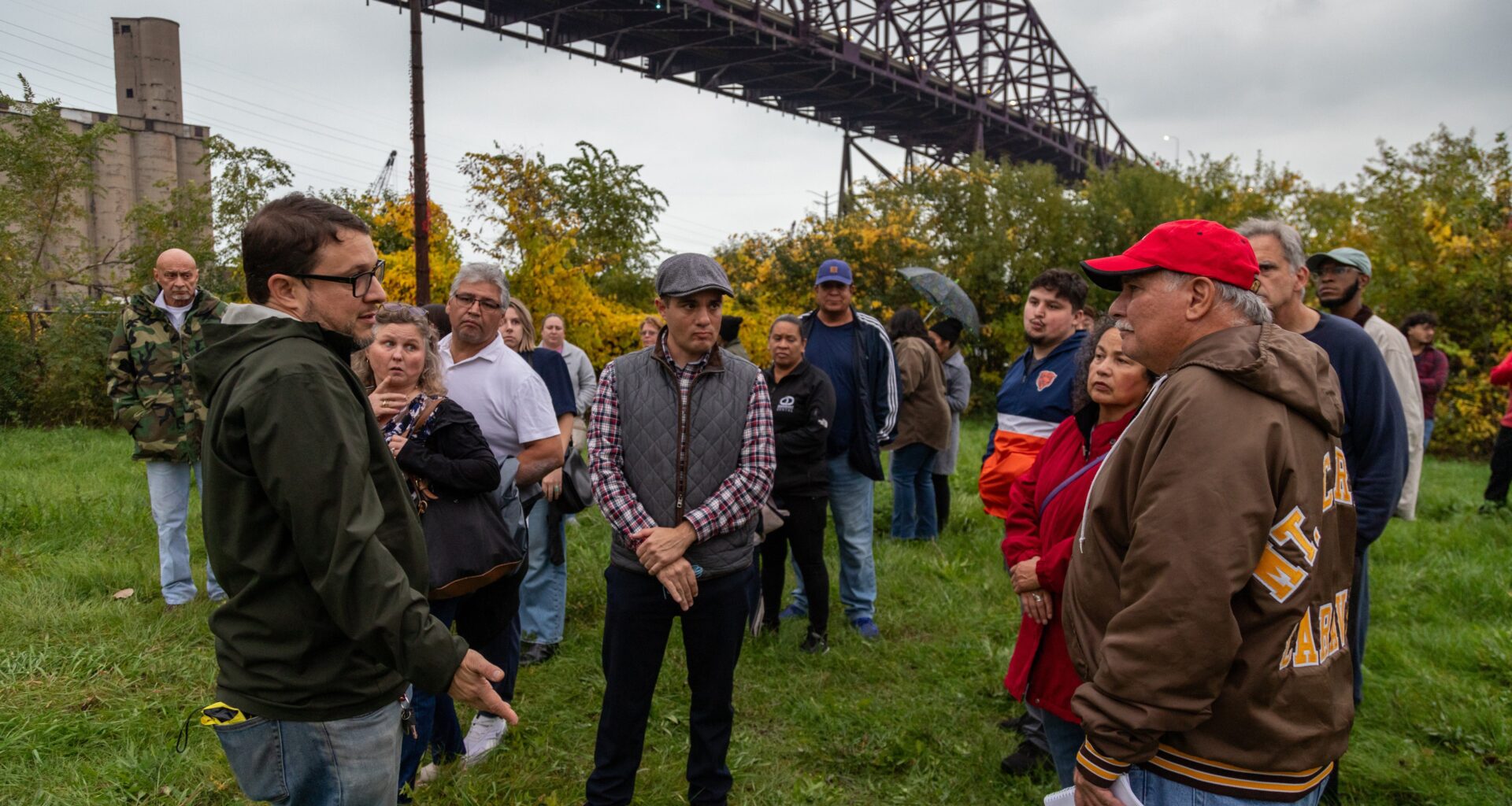 people stand and talk in a field under a bridge