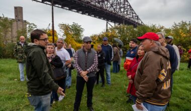 people stand and talk in a field under a bridge