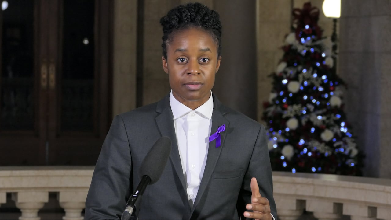 Person with dark hair and dark eyes wearing a suit blazer and white shirt stands at a podium with a microphone