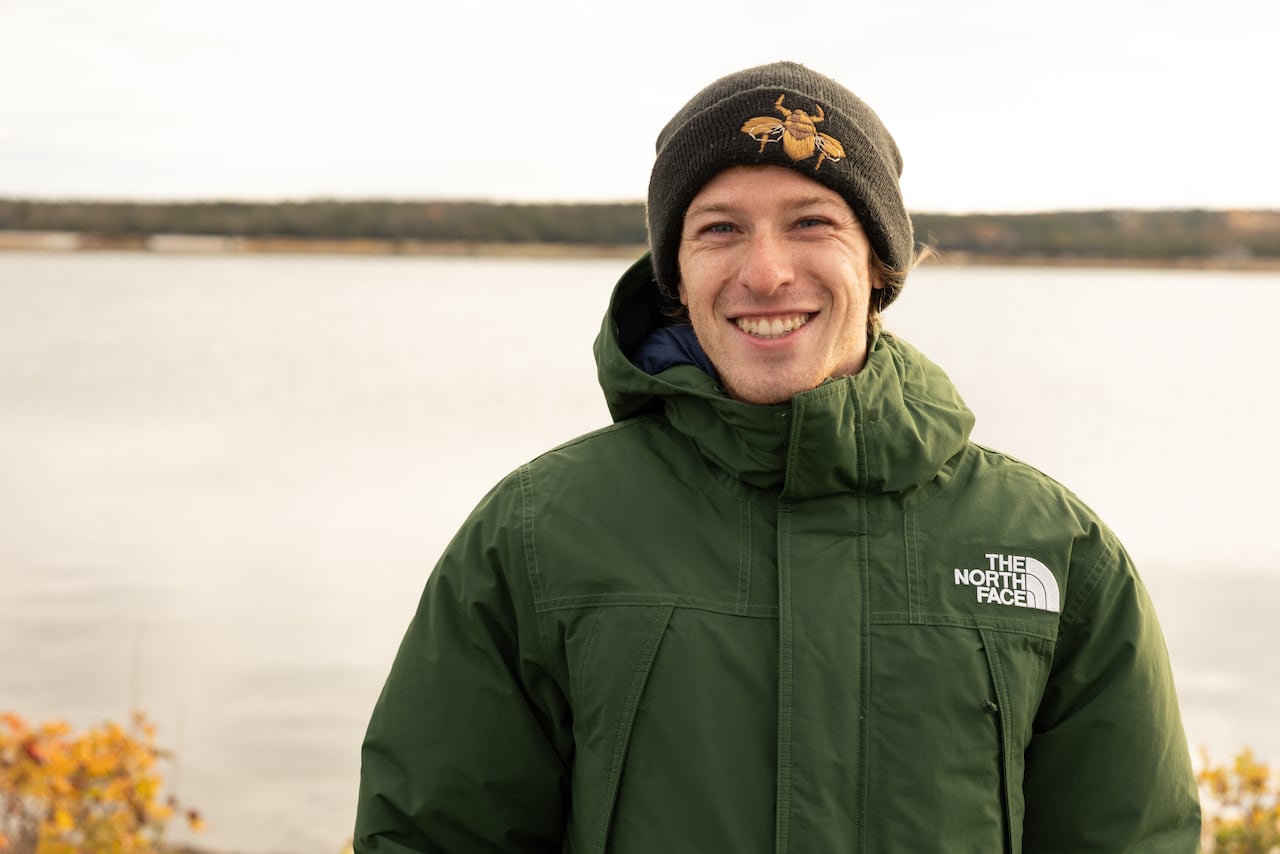 A man in a green coat and a tuque smiles. Water and a treeline are visible in the distance.