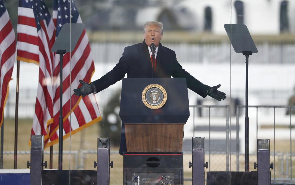 Donald Trump speaking at a rally with two American flags behind him.