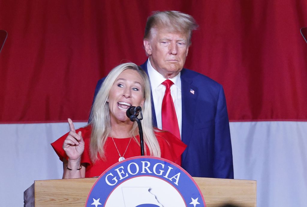 Marjorie Taylor Greene speaks at a podium with former President Donald Trump standing behind her.