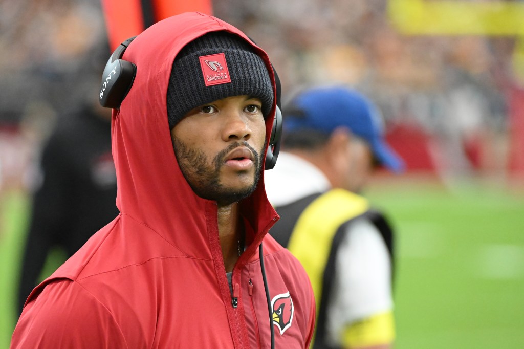 Arizona Cardinals player in red hoodie and black beanie, wearing headphones on the sideline.