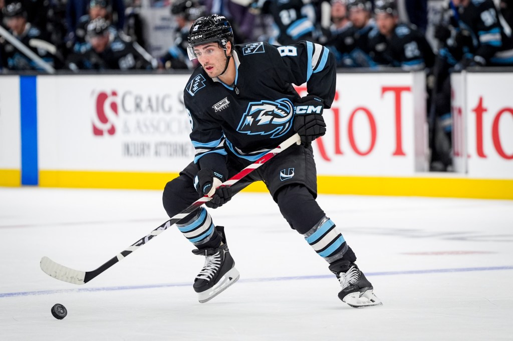 A Utah Mammoth hockey player in a black and light blue jersey skates with the puck on the ice.