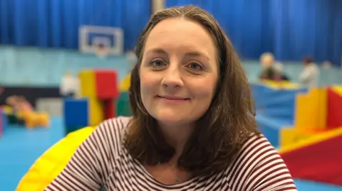 Camille Ross-Burrows - A woman with brown shoulder length hair, and a brown and white striped jumper. 