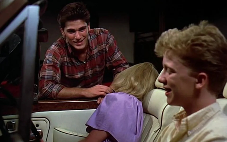 Two people are in a convertible car. One leans against the driver’s side talking to the smiling driver, while a passenger slumps over unconscious