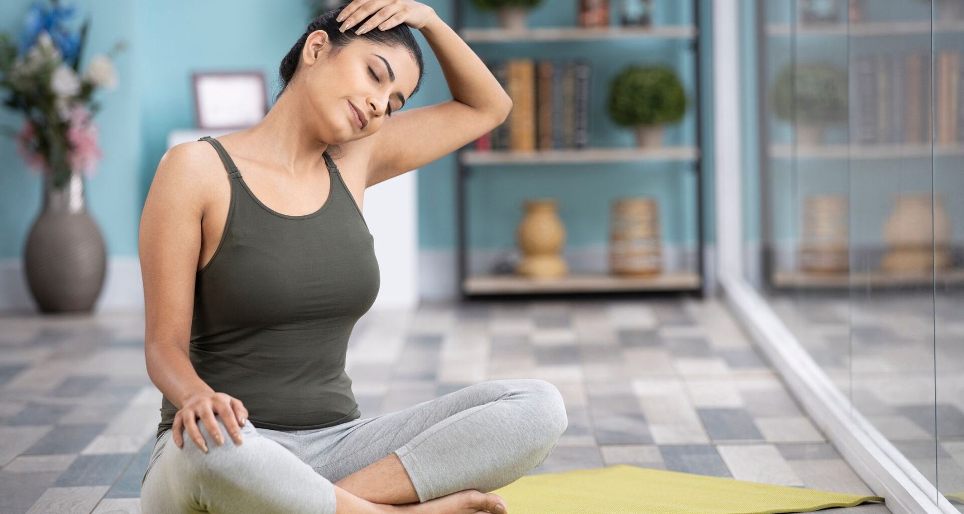 woman sitting crossed legged on a yellow mat on a grey lino floor tilting her head to the side and holding it with one hand, eyes closed