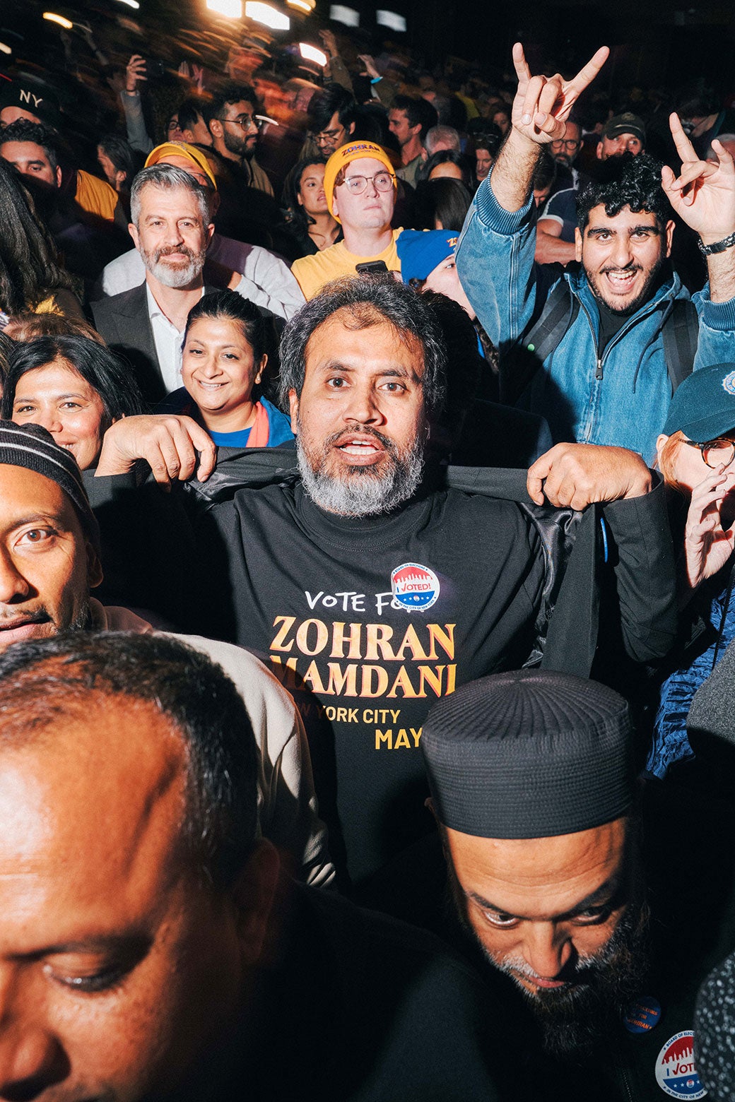 A man at the center of a crowd holds his jacket open so you can see his whole Vote for Zohran shirt. 