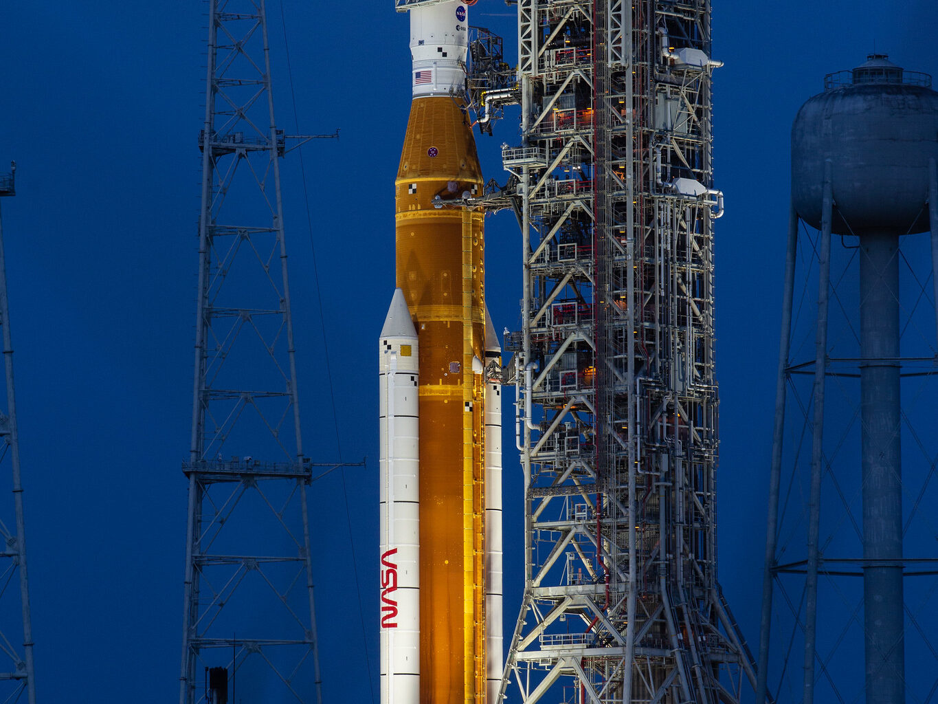 A large orange NASA rocket with white booster rockets stands vertically on a launch pad, lit by spotlights, with the full Moon and night sky behind it.