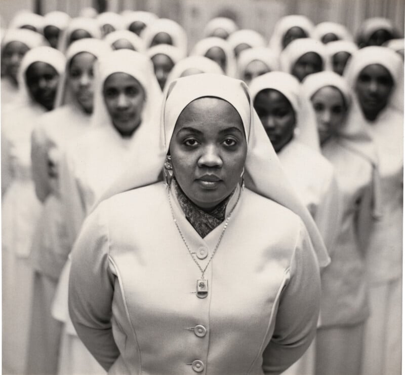 A group of women in matching white uniforms and head coverings stand in rows, with one woman at the front looking directly at the camera, wearing a necklace and pendant. The background women are slightly out of focus.