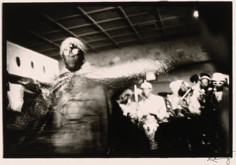 A person in a shiny costume and headpiece dances energetically on stage, with blurred motion. In the background, a band of musicians plays instruments in a dimly lit room.