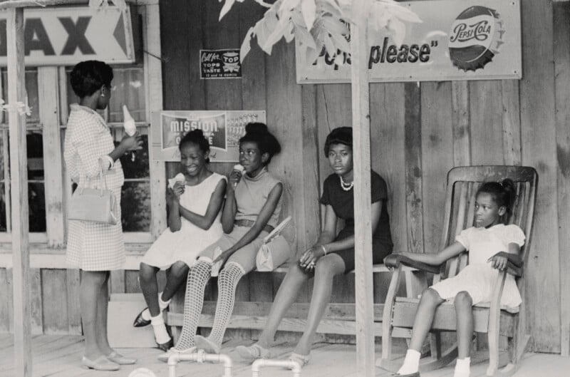 Five Black girls and women sit and stand on a wooden porch, eating ice cream. Four are seated, one stands holding a cone. Old signs, including Pepsi and TAX, hang on the building behind them.