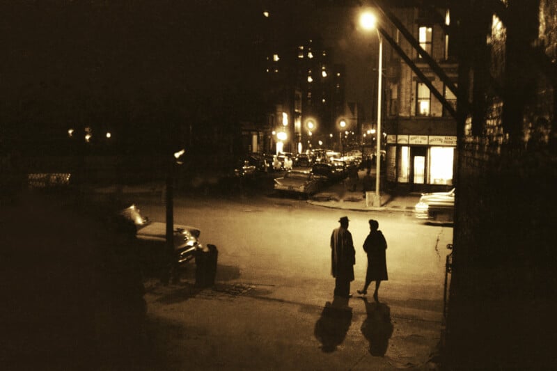 Two people in coats and hats stand on a dimly lit city street corner at night, illuminated by a streetlamp, with cars and buildings visible in the background.