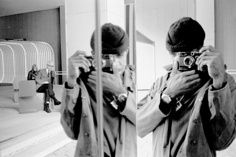 A person wearing a beanie takes a photo with a camera in front of a reflective surface, creating a mirrored image. In the background, two people sit on a bench inside a modern building.