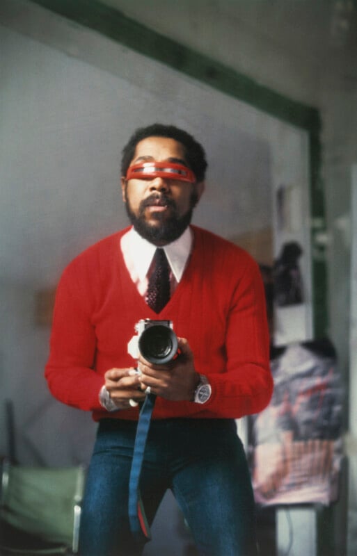 A bearded man wearing a red sweater, tie, and glasses holds a camera, looking into a mirror. The background shows a blurred room with a green-framed doorway and some indistinct objects.