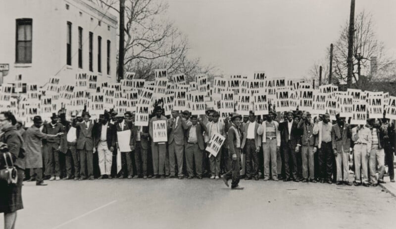 A large group of protesters, mostly men, stand in a line holding signs that read “I AM A MAN” during a civil rights demonstration on a city street. Some trees and a building are visible in the background.